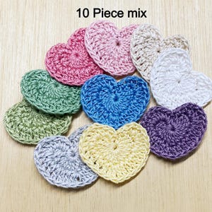 May include: Ten crocheted heart-shaped decorations in various colors, including pink, green, blue, yellow, purple, and white. The hearts are arranged in a semi-circle on a wooden surface. 10 Piece mix.