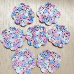 May include: Seven handmade crochet flower coasters in pastel colors. Each coaster features a floral design with a mix of pink, blue, purple, and cream yarn. The coasters are arranged on a light brown wooden surface.