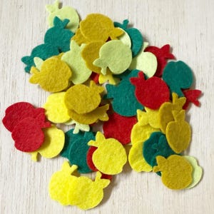 May include: A collection of small, flat, die-cut felt shapes in the form of apples. The pieces are rendered in a vibrant palette of red, yellow, lime green, and dark teal, featuring a simple silhouette with a small stem at the top of each.