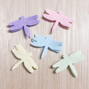 May include: Five felt dragonfly shapes in pastel colors: purple, pink, light blue, yellow, and green. The dragonflies are cut out and have a simple design.