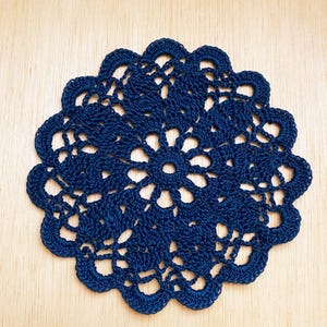 May include: A navy blue, handmade crochet doily. The circular doily features an intricate openwork design with a scalloped edge. The detailed pattern is visible against the light wood surface.