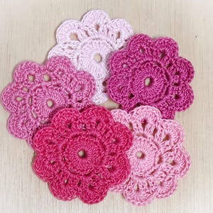 May include: Five circular crochet flower appliques arranged in a cluster. The pieces feature a central hole and scalloped edges, crafted in varying shades of pink, ranging from a pale pastel pink to a deep, vibrant magenta and rose pink.