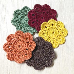May include: Five circular crochet coasters in various colors including sage green, deep burgundy, bright yellow, burnt orange, and dark brown. Each coaster features a central circular pattern with a scalloped, flower-like edge design.