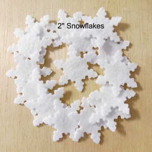 May include: A collection of white felt snowflakes, each measuring 2 inches, arranged on a light brown surface. The snowflakes are in a circular pattern. The text "2" Snowflakes" is printed in black.