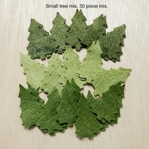 May include: A collection of 30 small, felt tree cutouts in various shades of green. The trees are arranged on a light-colored wooden surface. Text at the top reads "Small tree mix, 30 piece mix."