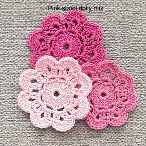 May include: Three handmade crochet flower doilies in shades of pink. The doilies are arranged on a gray textured surface. The text "Pink spool doily mix" is visible at the top of the image. The doilies are circular with a floral design.
