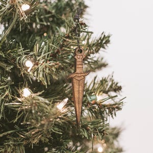 Peut inclure: Une décoration d'épée en bois suspendue à un sapin de Noël. La décoration est brune et a un design simple.