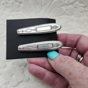 May include: Two silver-toned hair clips featuring an elongated, tapered shape with an ornate, embossed pattern. Each clip has a central rectangular panel and intricate decorative detailing along the edges, mounted on a black rectangular card.