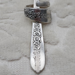 May include: A silver-toned metal ring crafted from a bent spoon handle. The ring features an embossed floral pattern with detailed roses and leaves along the band. The metal has a polished finish with visible oxidation in the recessed areas.