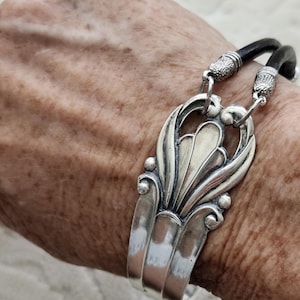 May include: A bracelet featuring a silver-toned metal centerpiece with an ornate, embossed fan-like design. The centerpiece is attached to two black leather cords by textured silver-toned metal end caps, creating a layered, flexible band.