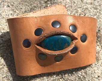 May include: A brown leather cuff bracelet with a turquoise stone set in the center. The bracelet is decorated with silver studs around the stone.
