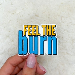 May include: A sticker with the words "FEEL THE BURN" in bold, block letters. The text is in a blue and yellow color scheme, with a white outline. The sticker is held by a hand against a white, textured background.