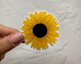 May include: A yellow sunflower sticker with a black center. The sticker has a white border and is held against a white background. The sunflower petals are detailed with yellow and orange hues.