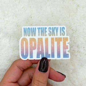 以下が含まれることがあります： 「NOW THE SKY IS OPALITE」の文字が青、オレンジ、ピンクのグラデーションで描かれたステッカー。白いテクスチャの背景に貼られています。