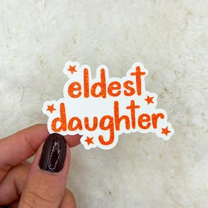 May include: A white sticker with the words "eldest daughter" in orange glitter lettering. The sticker has a white border and orange star accents. The sticker is held by a hand with dark nail polish.