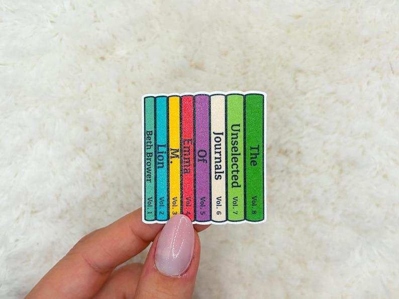 May include: A sticker featuring a stack of colorful book spines. The spines are labeled with titles like "Beth Brower," "Lion," "Emma," and "The Unselected Journals." Each spine is a different color, including teal, yellow, red, purple, and green.