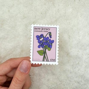 Può includere: Un francobollo vintage con sfondo rosa che presenta un disegno a linea di un fiore di violetta comune viola. Il francobollo riporta la scritta "new jersey common violet 1788".