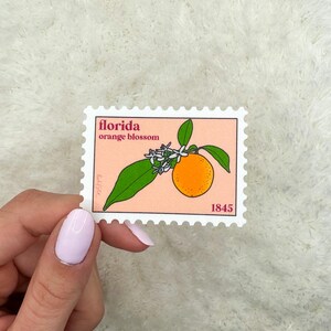 May include: A vintage-style stamp with a white border and a pink background. The stamp features a drawing of an orange blossom with a single orange fruit. The text "florida orange blossom" and "1845" are printed on the stamp.