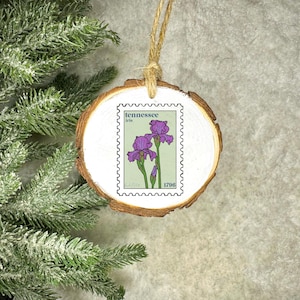 May include: A round wooden ornament with a Tennessee Iris stamp design. The stamp features purple irises on a light green background. The ornament has a natural wood grain edge and a twine loop for hanging.