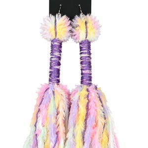 May include: A pair of long drop earrings featuring a purple yarn-wrapped post topped with a fluffy pastel pom-pom. The base consists of a large, multi-coloured tassel made from soft, feathery yarn in shades of pink, yellow, blue, and lavender.