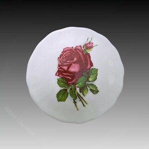 Puede incluir: Tapa de urna blanca y redonda con una ilustración detallada de una rosa roja oscura con hojas verdes y un capullo. El diseño floral está centrado en la tapa, que tiene un borde ligeramente irregular. Es un artículo decorativo.