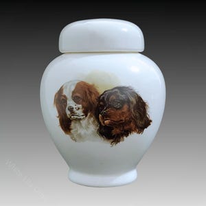 Op de afbeelding: Witte keramische urn met een deksel, met een geschilderd beeld van twee Cavalier King Charles Spaniëls. De honden kijken naar voren met hun hoofden lichtjes naar de zijkant gekanteld. De ene hond is bruin en wit, de andere is zwart en bruin.