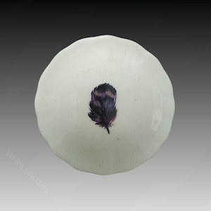 Puede incluir: Un pequeño plato de cerámica blanca con un borde festoneado. El plato tiene un diseño de una sola pluma morada en el centro.