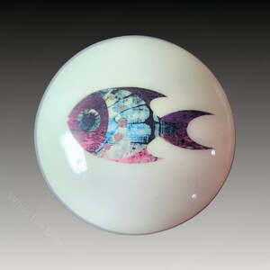 Puede incluir: Un adorno redondo de cerámica blanca con un diseño de pez colorido. El pez es de color morado, rosa, azul y blanco con un contorno negro.
