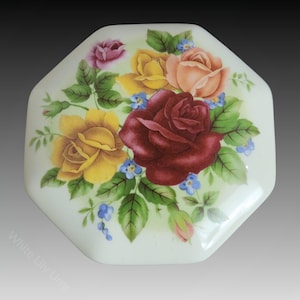 Puede incluir: Una tapa octogonal de cerámica blanca con un arreglo floral central de rosas de color rojo oscuro, amarillo y melocotón rodeadas de hojas verdes y pequeñas flores azules. El texto White Lily Urns está impreso en la esquina inferior izquierda.