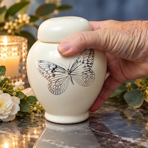 Puede incluir: Una urna de cerámica de color crema con tapa redondeada y cuerpo ancho, que presenta una ilustración detallada de una mariposa plateada en la parte frontal. El recipiente tiene un acabado liso y brillante con una forma clásica.