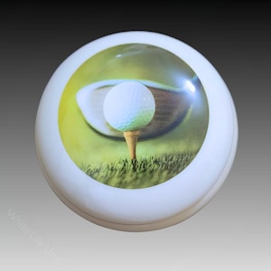 May include: A white, round urn with a golf-themed image. The image features a golf ball on a tee, a golf club, and green grass. The urn is a memorial product.