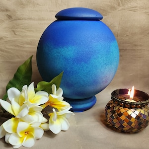 May include: A blue urn with a matching lid, a decorative candle holder with a lit candle, and white and yellow flowers with green leaves. The urn is spherical with a textured finish.