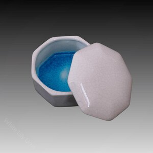 Puede incluir: Una caja de cerámica octogonal blanca con tapa. La caja está abierta y el interior es de color azul brillante.