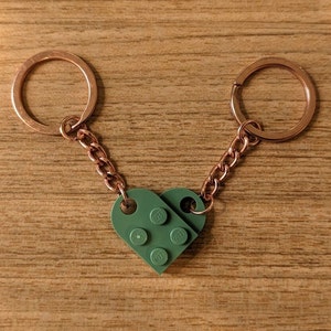 May include: A pair of keychains featuring a green heart made of Lego bricks. Each keychain has a copper-colored ring and chain. The heart is made of two Lego bricks connected together. The background is a wooden surface.