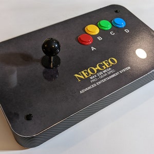 May include: Black and gray Neo Geo arcade controller with a joystick and colored buttons. The controller has the text "NEO-GEO" and "MAX 330 MEGA PRO-GEAR SPEC ADVANCED ENTERTAINMENT SYSTEM". The buttons are red, yellow, green, and blue.