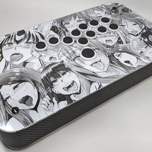 May include: A rectangular arcade controller featuring a black and white manga-style collage print on the top surface. The device includes multiple circular black buttons with white borders arranged in a layout for gaming, with a dark carbon fiber texture.