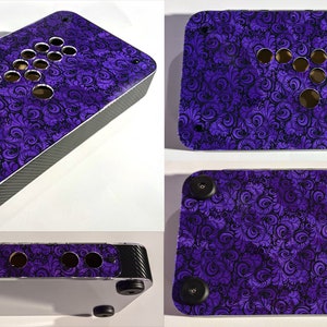 May include: A custom arcade controller with a purple and black floral design. The controller has multiple button holes and a joystick layout. The sides have a carbon fiber texture, and the bottom has black rubber feet.