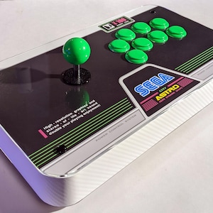 May include: A black and white Sega City Astro arcade controller with a green joystick and green buttons. The controller has the text "High-resolution graphics and stereo of the art sound double your playing enjoyment!"