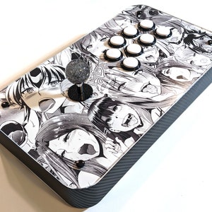 May include: A custom arcade stick featuring a black and white manga-style graphic design. The top panel has a printed design with multiple manga faces. It has eight white buttons and a joystick with a gray ball top.