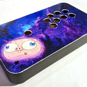 May include: Custom arcade game controller with a vibrant galaxy design and a cartoon pig character. The controller features a carbon fiber finish on the sides and a layout for buttons and a joystick. The top panel showcases a colorful nebula.
