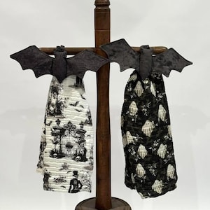 May include: Two hanging kitchen towels with dark gray fabric bat-shaped toppers. One towel features a white background with a black gothic-style print of gates, skulls, and figures. The second towel has a black background with a white ghost pattern.