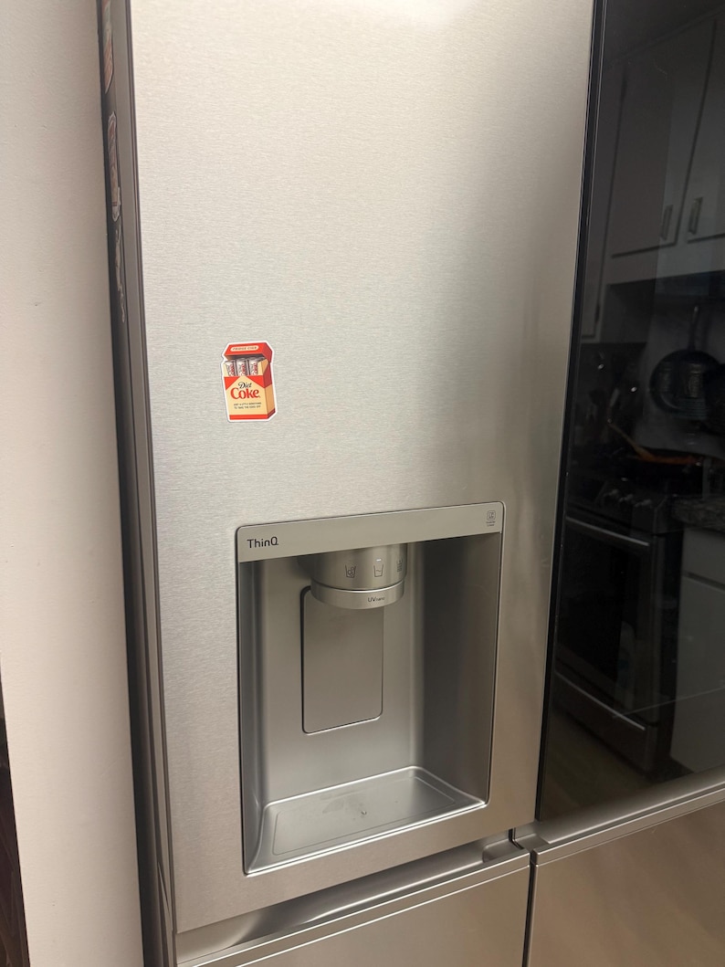 May include: Stainless steel refrigerator with a water and ice dispenser. A small Coca-Cola sticker is on the door. The refrigerator has a sleek, modern design with a black glass door on the right side.