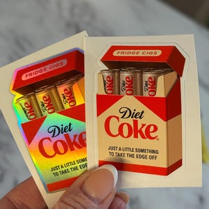 K&ouml;nnte beinhalten: Zwei Aufkleber mit einem Diet Coke-Box-Design, mit dem Text "FRIDGE CIGS". Jede Box enth&auml;lt drei Diet Coke-Dosen. Der Text "Diet Coke" wird zusammen mit dem Satz "JUST A LITTLE SOMETHING TO TAKE THE EDGE OFF" angezeigt.