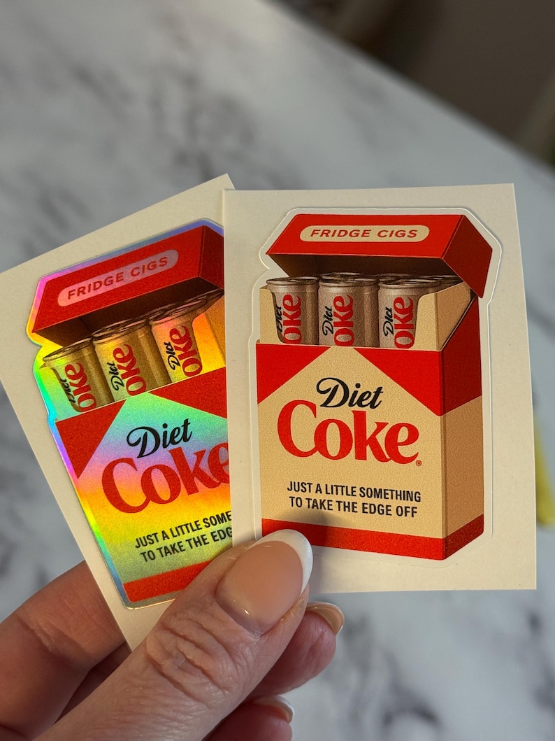 K&ouml;nnte beinhalten: Zwei Aufkleber mit einem Diet Coke-Box-Design, mit dem Text "FRIDGE CIGS". Jede Box enth&auml;lt drei Diet Coke-Dosen. Der Text "Diet Coke" wird zusammen mit dem Satz "JUST A LITTLE SOMETHING TO TAKE THE EDGE OFF" angezeigt.