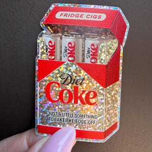 May include: A holographic sticker shaped like a cigarette pack with red accents. The pack features the text FRIDGE CIGS at the top, Diet Coke branding, and the phrase JUST A LITTLE SOMETHING TO TAKE THE EDGE OFF. Three soda cans are visible inside.