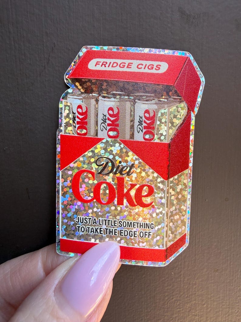 May include: A holographic sticker shaped like a cigarette pack with red accents. The pack features the text FRIDGE CIGS at the top, Diet Coke branding, and the phrase JUST A LITTLE SOMETHING TO TAKE THE EDGE OFF. Three soda cans are visible inside.