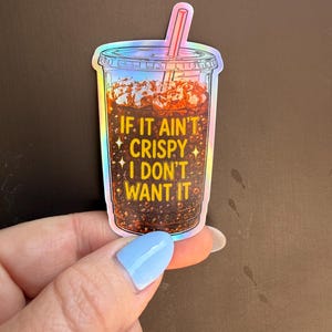 May include: A holographic sticker shaped like a clear plastic cup filled with a dark brown carbonated beverage, ice cubes, and a pink straw. The text IF IT AIN'T CRISPY I DON'T WANT IT is printed in yellow letters on the front of the cup.