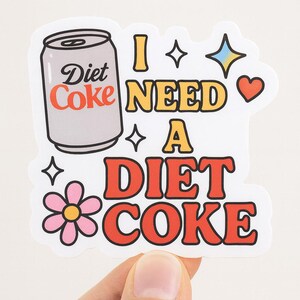 May include: A white sticker with the words "I NEED A DIET COKE" in a retro font. The sticker features a Diet Coke can graphic, stars, a heart, and a flower.