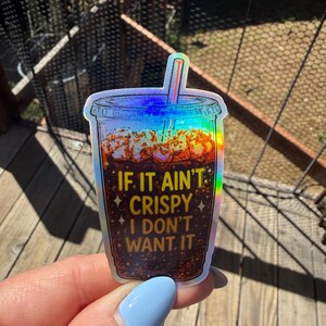 May include: A holographic sticker shaped like a plastic cup filled with a dark brown carbonated beverage and ice. The text IF IT AIN'T CRISPY I DON'T WANT IT is printed in yellow on the front of the cup, accompanied by small star icons.