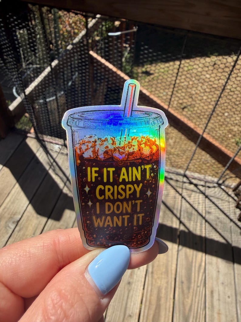 May include: A holographic sticker shaped like a plastic cup filled with a dark brown carbonated beverage and ice. The text IF IT AIN'T CRISPY I DON'T WANT IT is printed in yellow on the front of the cup, accompanied by small star icons.